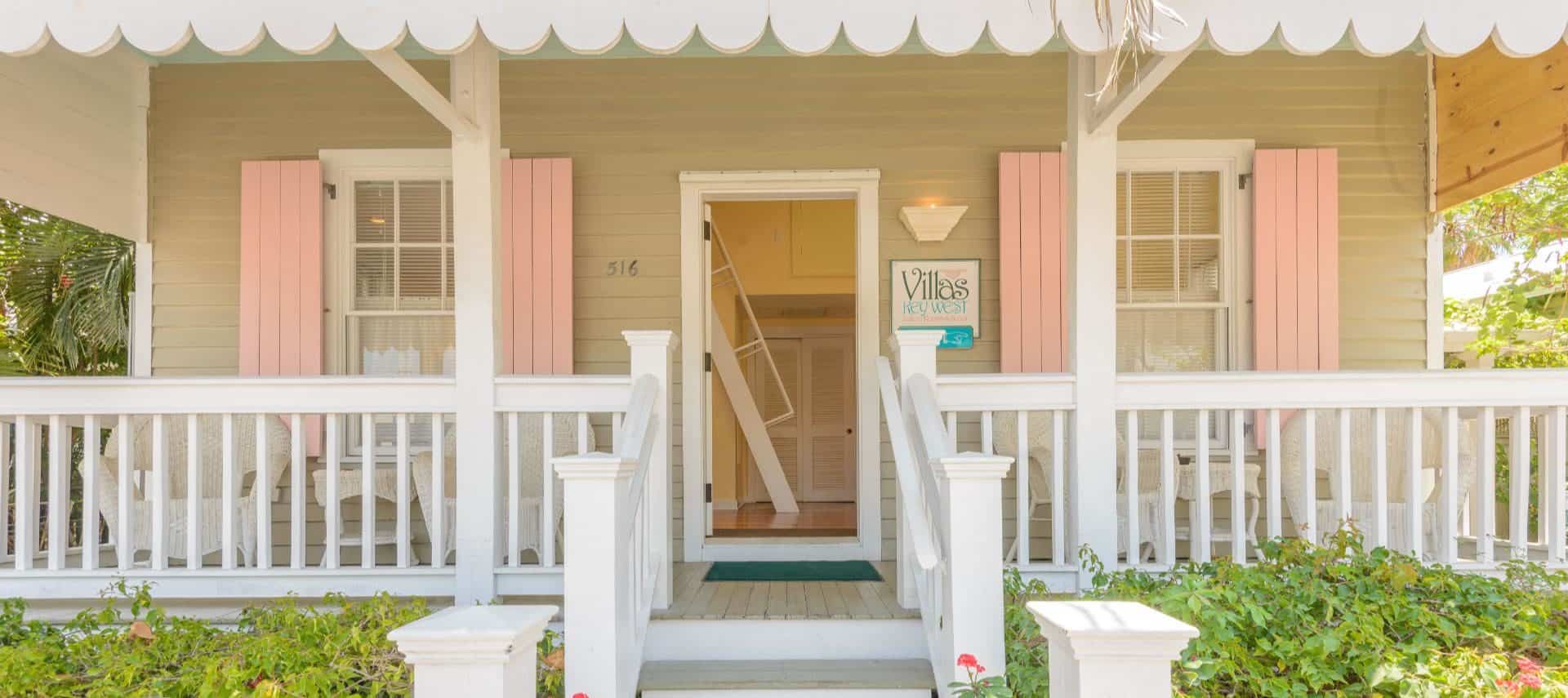 Front entrance of villa painted light tan with white trim and porch and pink shutters surrounded by lush greenery