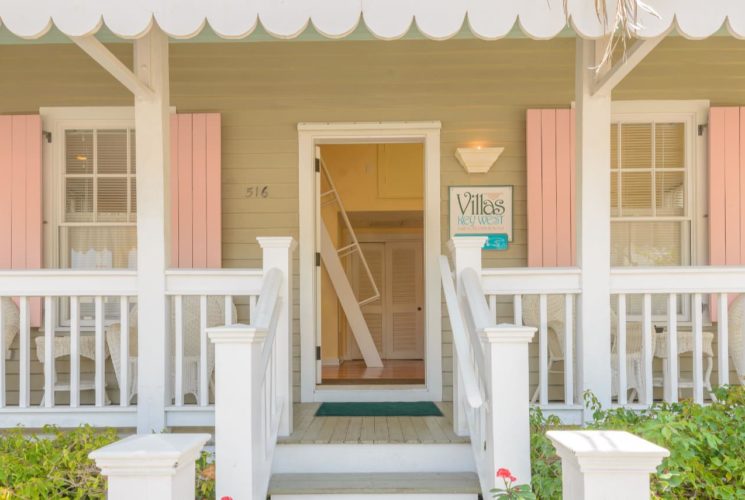 Front entrance of villa painted light tan with white trim and porch and pink shutters surrounded by lush greenery